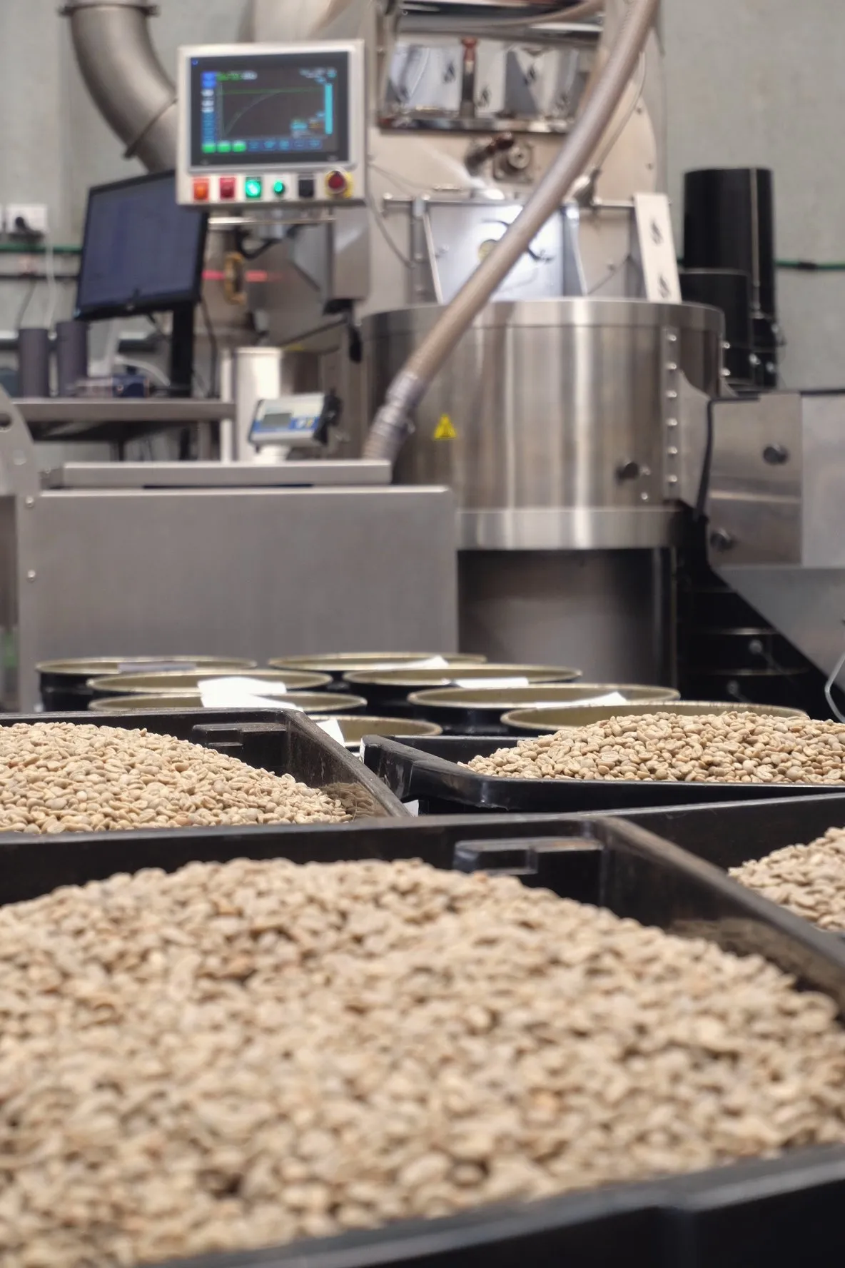A roaster's digital control panel behind bins of green coffee